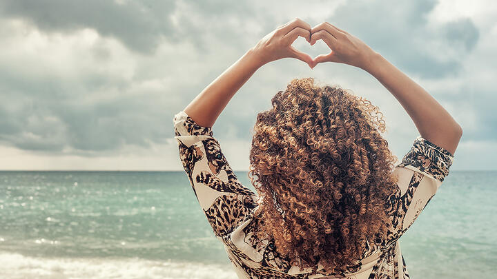 A woman with beautiful curly hair at the beach, forming a heart with her fingers above her head